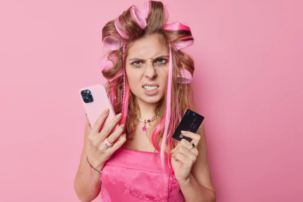 אשה צעירה לבושה ורוד, על רקע ורוד. רולים בשיער, מחזיקה טלפון וכרטיס אשראי, מתוסכלת