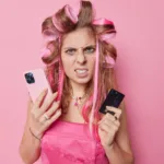 אשה צעירה לבושה ורוד, על רקע ורוד. רולים בשיער, מחזיקה טלפון וכרטיס אשראי, מתוסכלת
