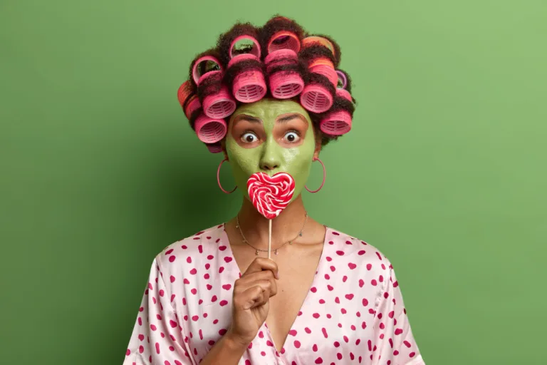 אשה עם קרם פנים ירוק ורולים בשיער אוחזת בסוכריה על מקל