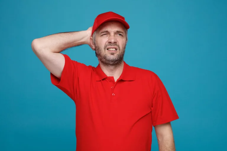 גבר עם כובע מצחיה אדום וחולצה אדומה ניצב על רקע קיר כחול במבט מבולבל